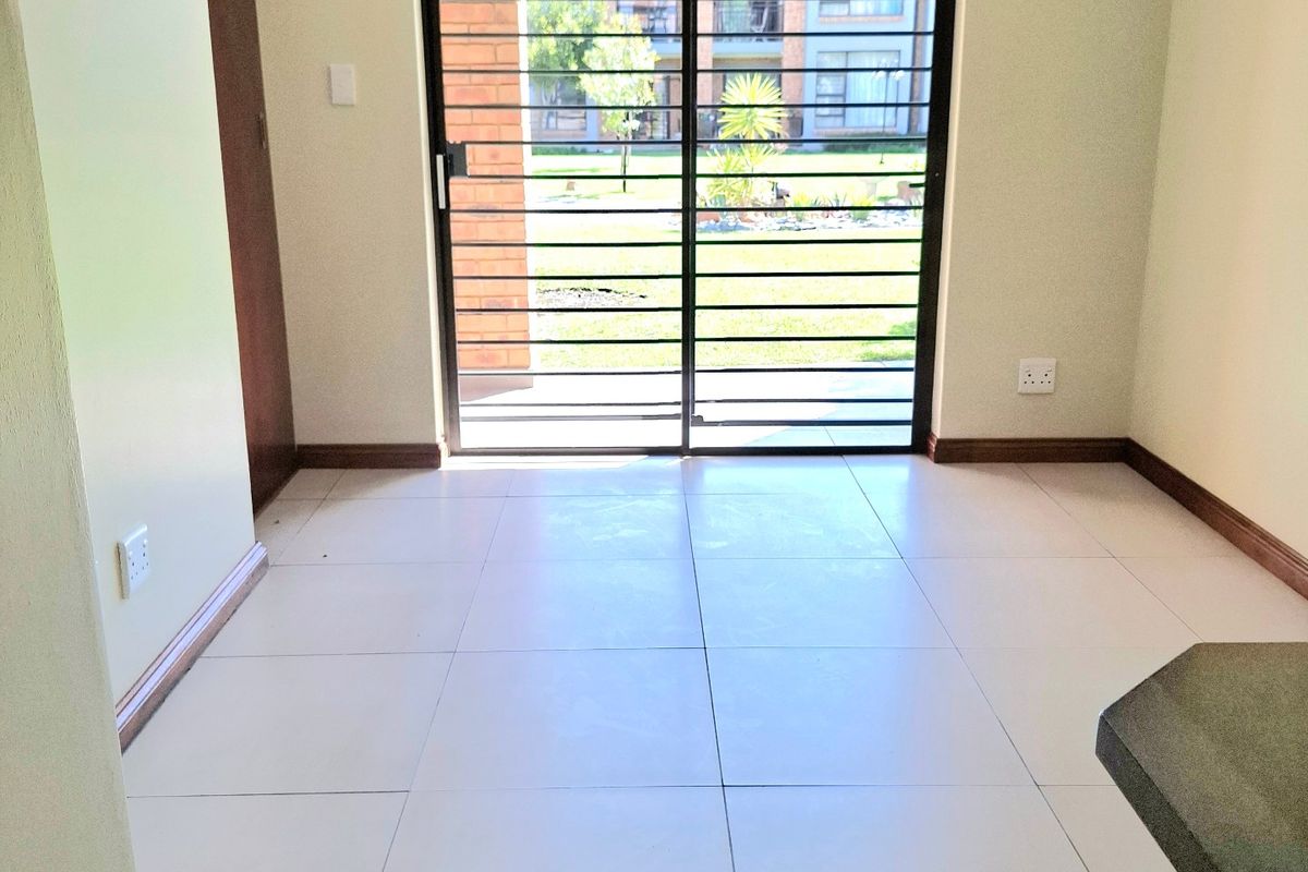 An empty, well-lit living area featuring neutral tiled flooring and a sliding glass door with security gate leading to the exterior.