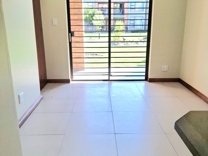 An empty, well-lit living area featuring neutral tiled flooring and a sliding glass door with security gate leading to the exterior.