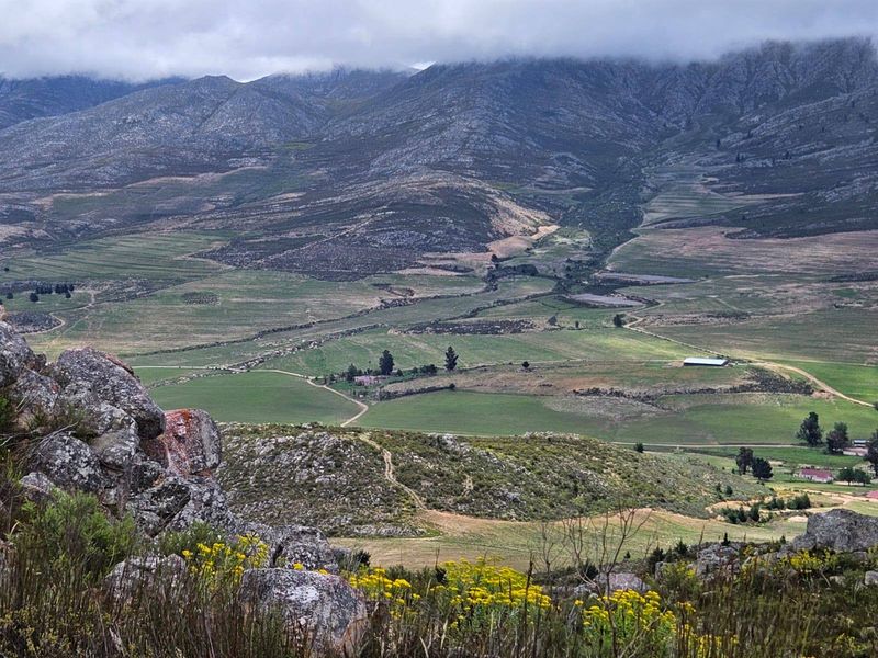 A scenic elevated landscape view overlooking expansive farm fields, rolling hills, and dramatic mountain ranges under a cloudy sky.