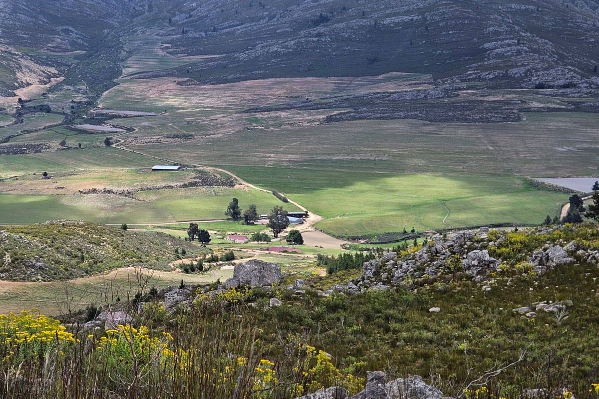 An elevated aerial view of a rural farm property set within a vast, mountainous landscape featuring open fields and farm infrastructure.