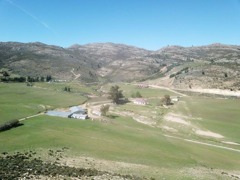 An aerial view of a large farm property featuring multiple outbuildings, open grazing land, and a dam set against a mountainous landscape.