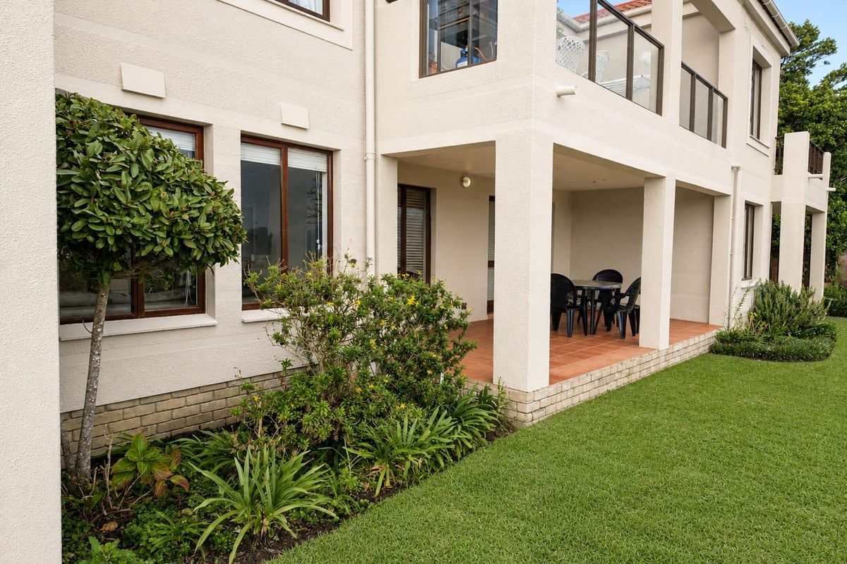 A well-maintained rear exterior view of the apartment building featuring a covered ground-floor patio, an upper-level balcony with glass balustrades, and a landscaped garden.