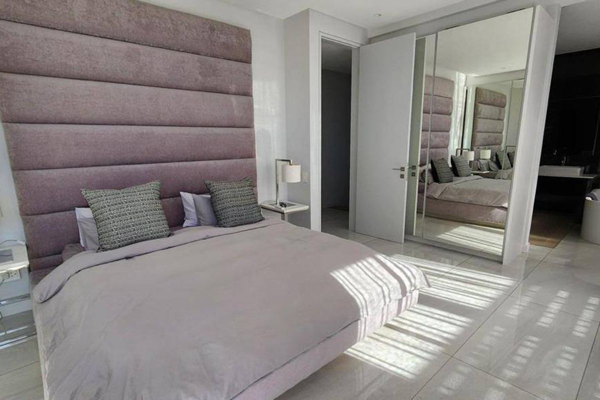 Modern bedroom featuring a large upholstered headboard, mirrored built-in wardrobes, tiled flooring, and an open-plan en-suite bathroom with a soaking tub.