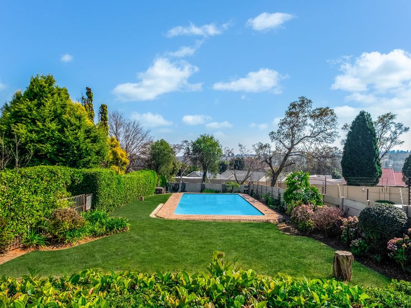 A well-maintained outdoor pool area featuring a clear swimming pool with brick coping, surrounded by a lush green lawn and established garden landscaping.