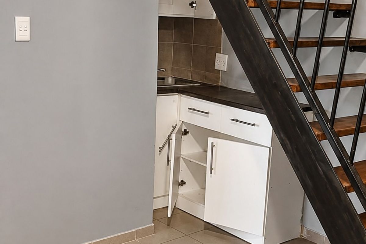 Compact kitchen area tucked under a staircase, featuring tiled flooring, built-in cabinetry, and exposed timber ceiling beams.