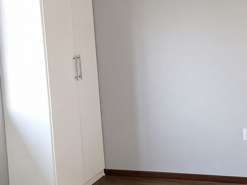 A clean, empty bedroom featuring modern laminate flooring, neutral grey walls, and a white built-in wardrobe with sleek handles.