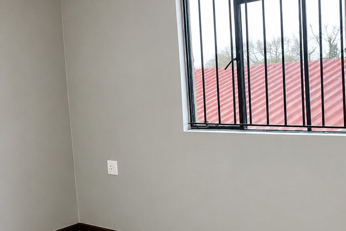 A clean, empty bedroom featuring modern laminate flooring, neutral grey walls, and a window fitted with security bars.