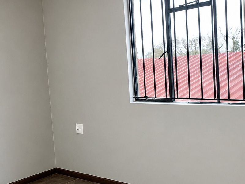 A clean, empty bedroom featuring modern laminate flooring, neutral grey walls, and a window fitted with security bars.