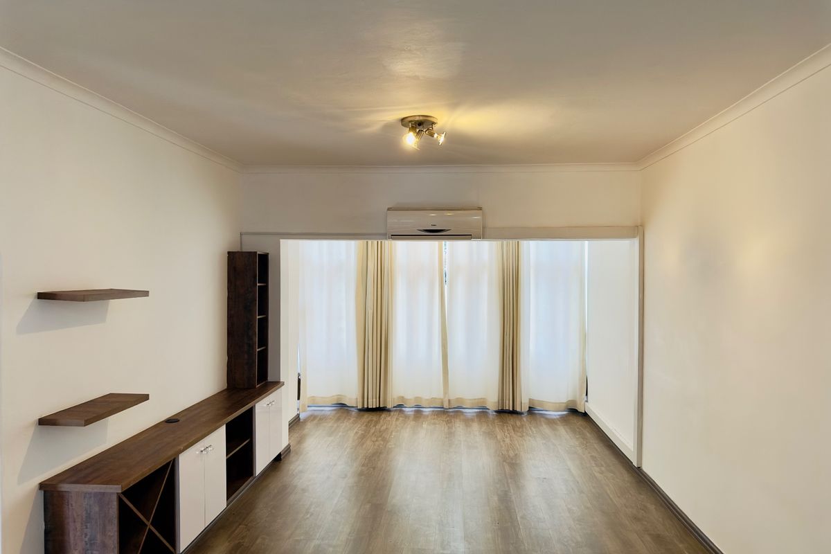 A bright, empty living room featuring wood-look laminate flooring, built-in cabinetry with shelving, and a wall-mounted air conditioning unit.