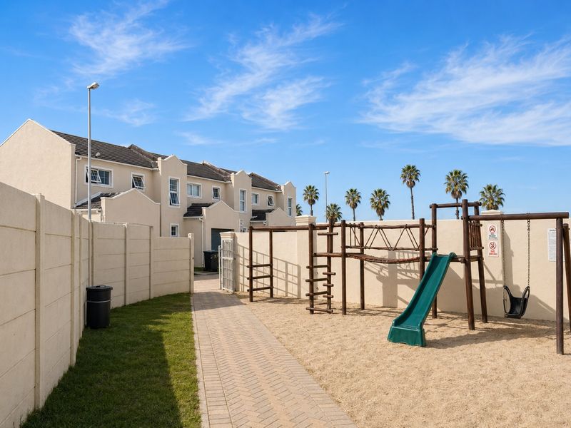 A well-maintained communal outdoor area featuring a paved walkway, sand-based playground with climbing frame and swing set, enclosed by precast concrete boundary walls.