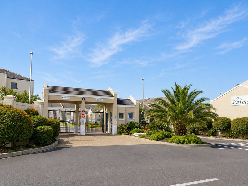 The secure entrance gate to The Palms apartment complex, featuring a paved driveway and well-maintained landscaping.