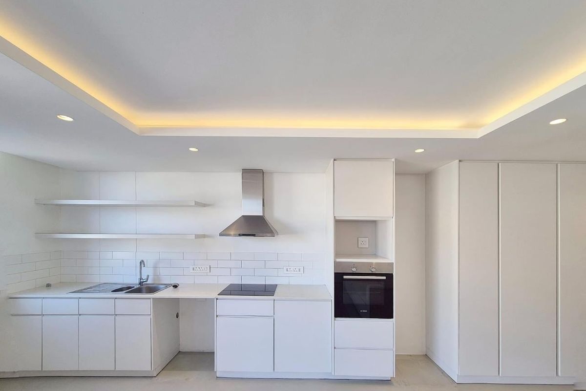 A modern, minimalist kitchen featuring white cabinetry, subway tile backsplash, integrated oven and hob, and elegant recessed cove lighting.