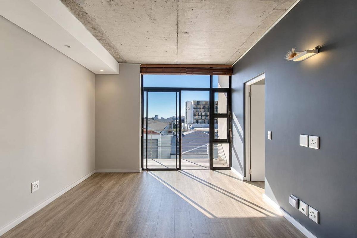 A bright, modern living area featuring an exposed concrete ceiling, laminate flooring, and a large sliding glass door leading to an outdoor balcony.