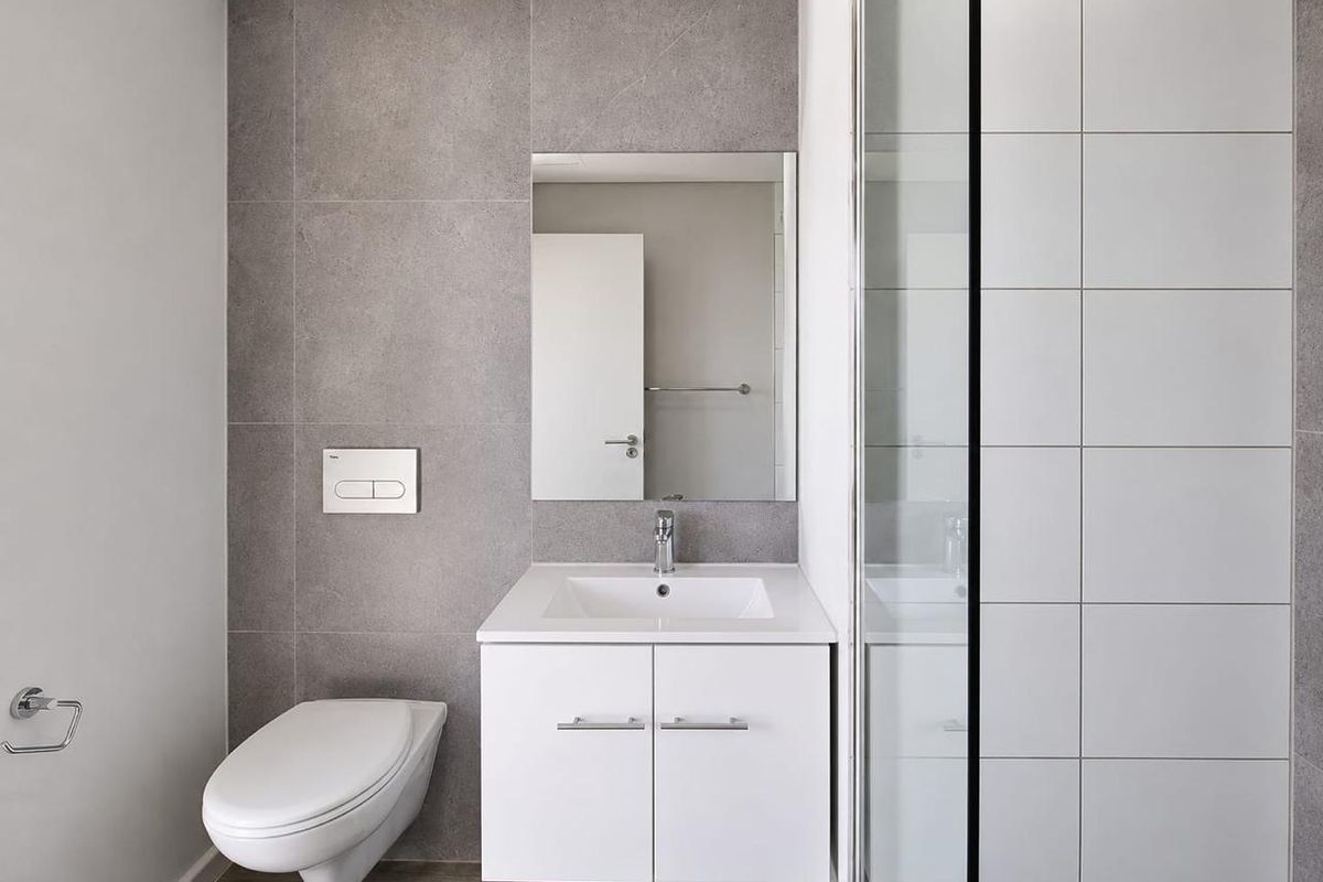 Modern bathroom featuring a wall-mounted toilet, sleek white vanity with integrated sink, and a glass-enclosed shower with contemporary grey and white tiling.