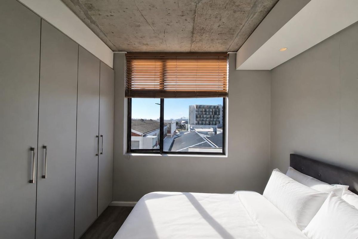 Modern bedroom featuring an exposed concrete ceiling, ample built-in cupboard storage, and a window with venetian blinds offering urban views.