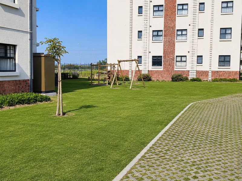 A well-maintained communal garden area featuring a manicured lawn, paved walkways, and playground equipment situated between apartment buildings.