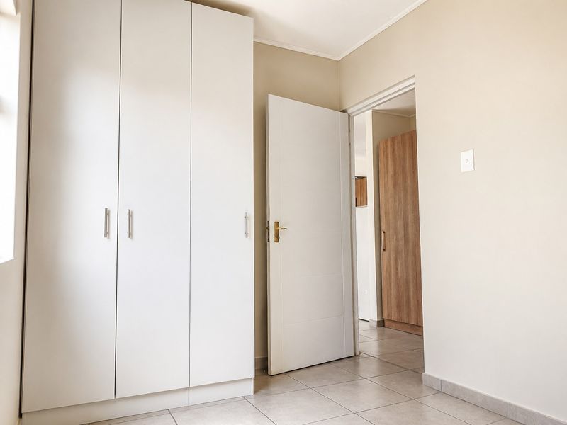 A bright, empty bedroom featuring neutral walls, tiled flooring, and a large built-in wardrobe with white cabinetry.