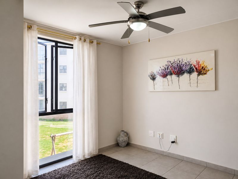 A bright and neutral bedroom featuring tiled flooring, a modern ceiling fan, and large windows overlooking the complex grounds.
