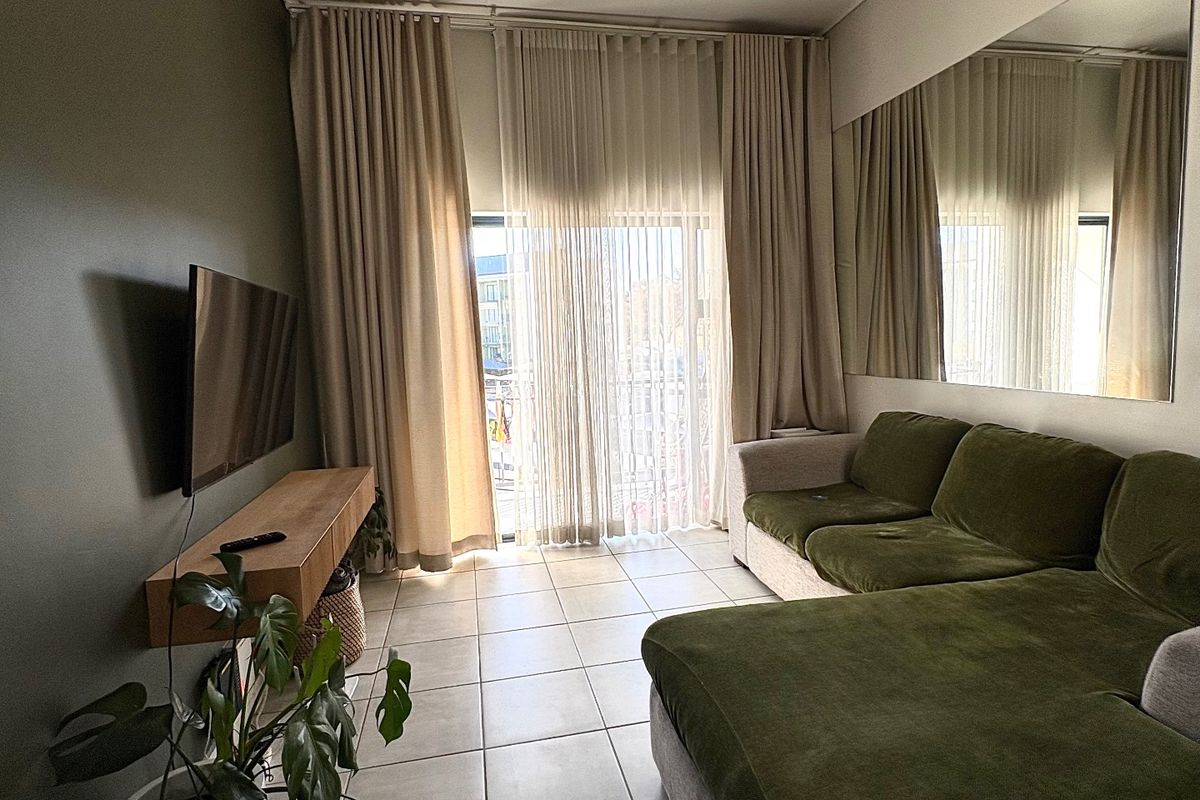 A well-maintained living room featuring tiled flooring, a wall-mounted shelf, and a large mirror, with natural light entering through floor-to-ceiling curtains.