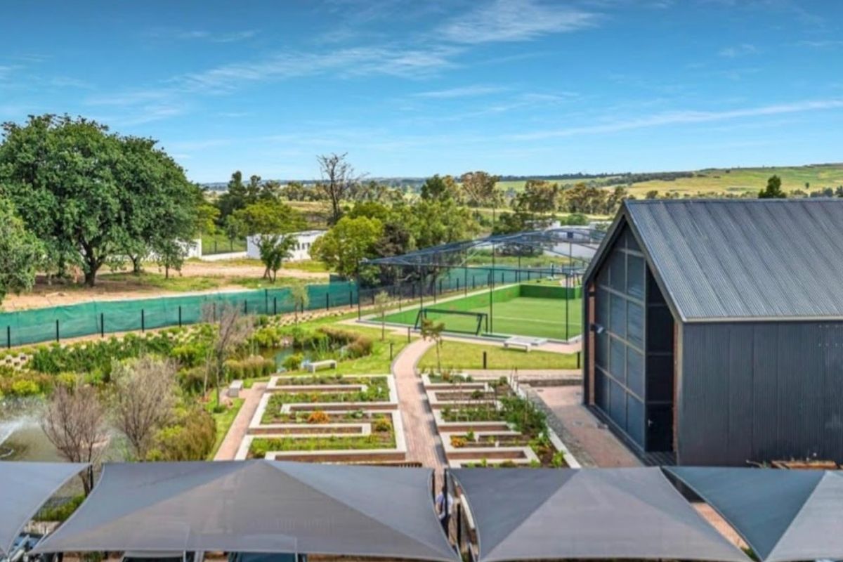 Elevated view of a well-maintained communal garden area featuring landscaped beds, a pond, a sports court, and a modern structure with a metal roof.
