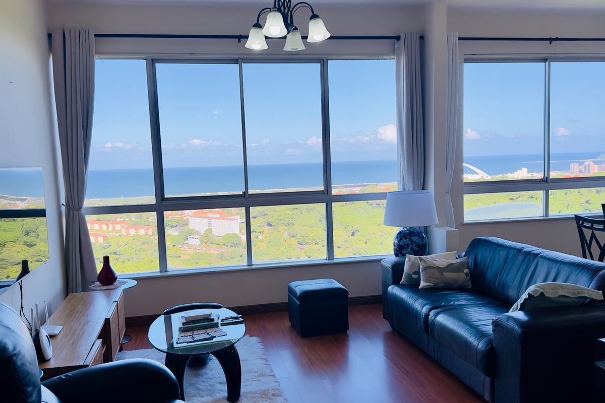 A bright living room featuring expansive floor-to-ceiling windows that offer panoramic coastal and landscape views, complemented by warm laminate flooring.