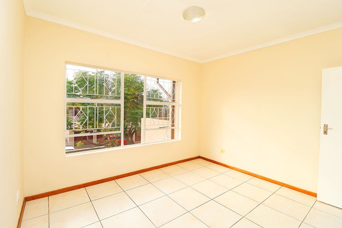 A bright, empty bedroom featuring neutral tiled flooring, large windows with security bars, and a simple ceiling light fixture.
