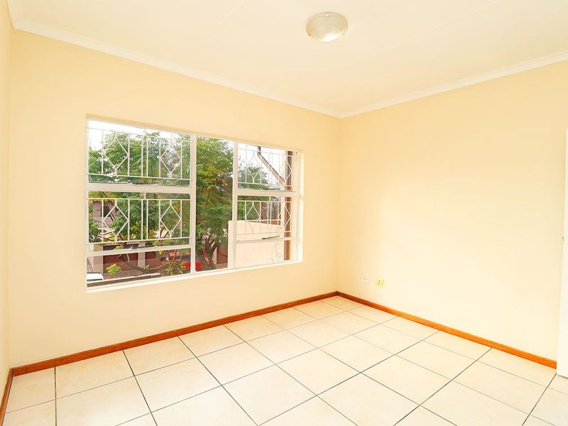 A bright, empty bedroom featuring neutral tiled flooring, large windows with security bars, and a simple ceiling light fixture.