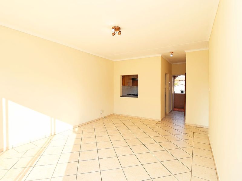 An empty, sunlit living room featuring neutral tiled flooring, coved cornicing, and a convenient pass-through window connecting to the kitchen area.
