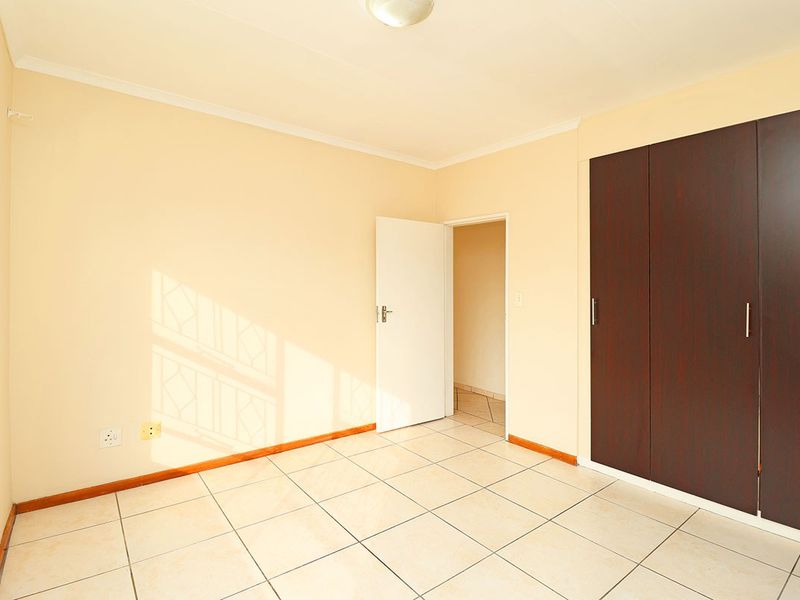 A bright, empty bedroom featuring neutral walls, tiled flooring, and a dark wood-finish built-in wardrobe with security bars on the window.