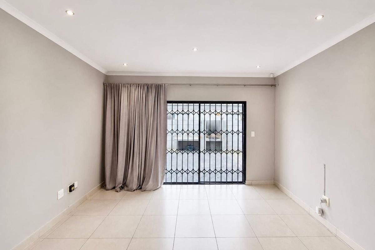 A bright, empty living room featuring neutral tiled flooring, recessed downlights, and a sliding door with a security gate leading to the exterior.