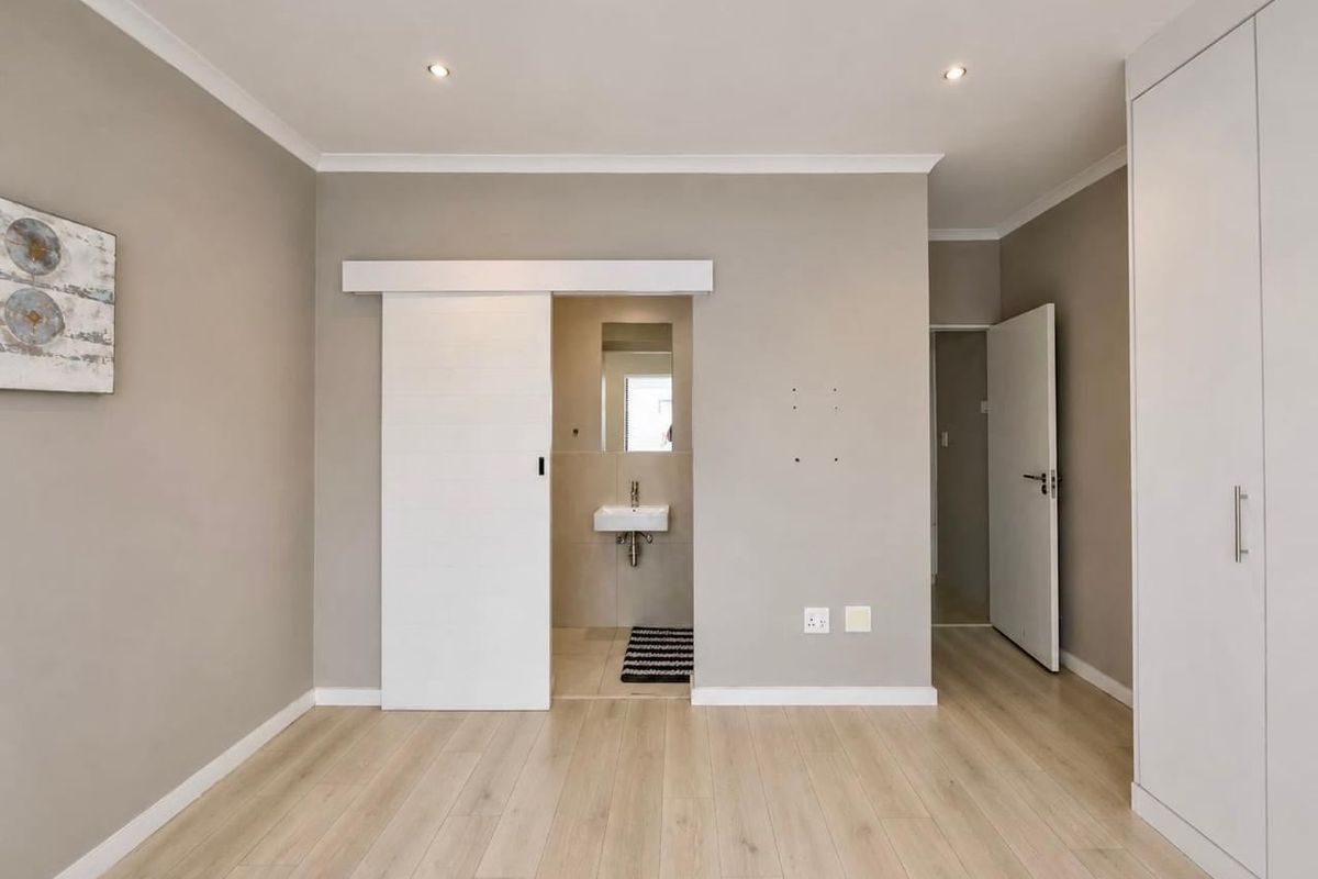 A clean, modern bedroom featuring light laminate flooring, built-in wardrobes, recessed lighting, and an en-suite bathroom accessed via a sliding door.