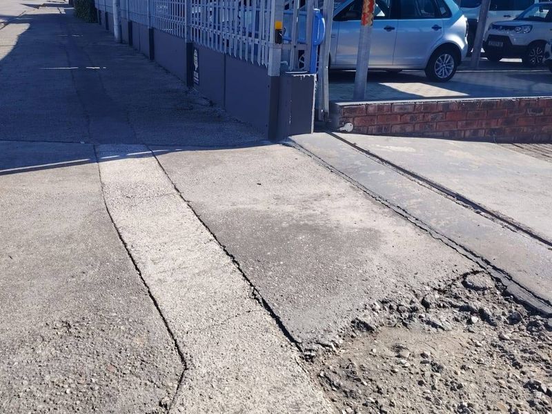 Street view of a commercial property entrance featuring steel palisade fencing and a shaded parking area with visible pavement damage at the driveway access.
