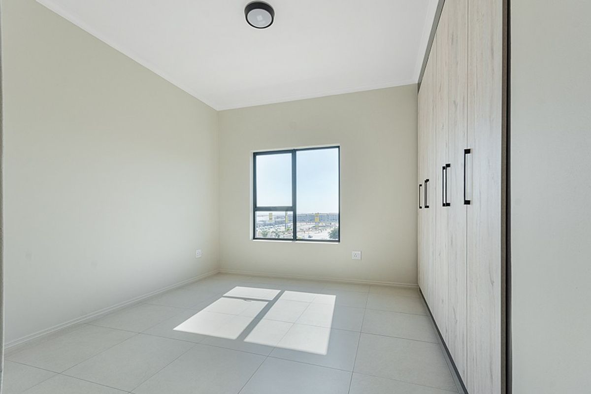 A bright, empty bedroom featuring modern built-in cupboards, neutral tiled flooring, and an aluminium-framed window with natural light.