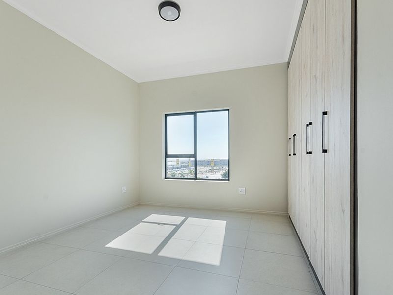 A bright, empty bedroom featuring modern built-in cupboards, neutral tiled flooring, and an aluminium-framed window with natural light.