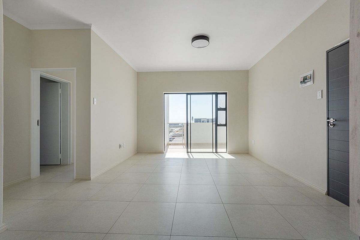 A bright, empty living room featuring neutral tiled flooring, a modern ceiling light, and a sliding glass door leading to an outdoor balcony area.
