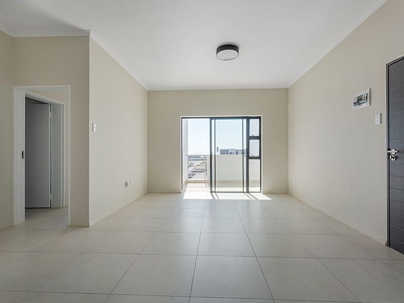 A bright, empty living room featuring neutral tiled flooring, a modern ceiling light, and a sliding glass door leading to an outdoor balcony area.
