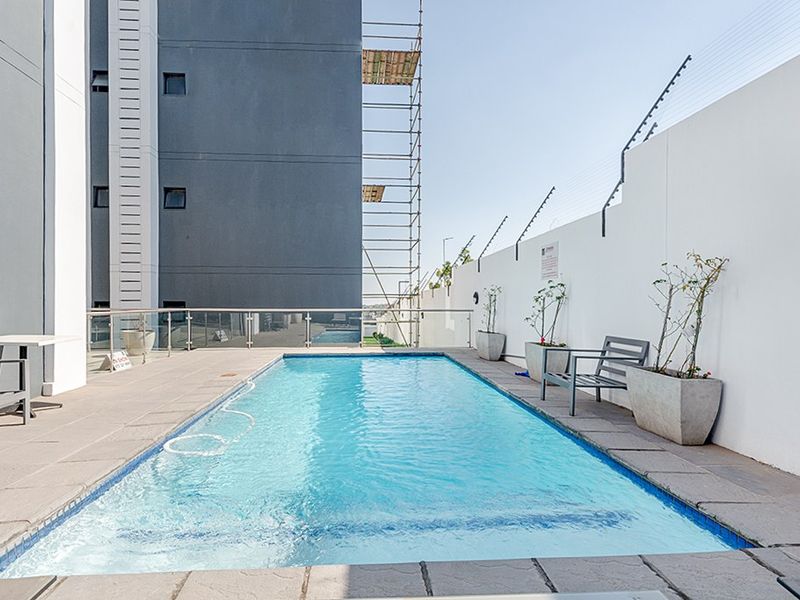 A clean, rectangular swimming pool area featuring a tiled deck, glass balustrade, and secure perimeter wall with electric fencing.