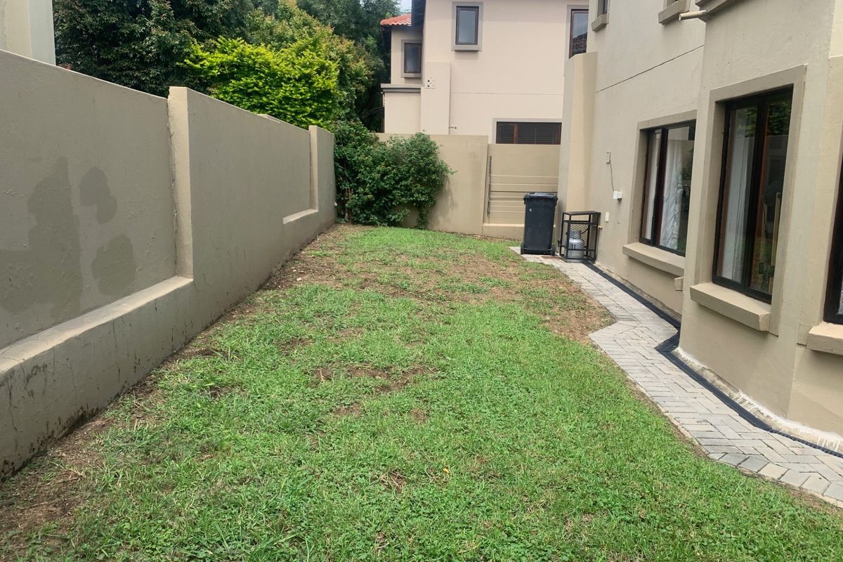 A rear garden area featuring a paved walkway alongside the building, a boundary wall, and a lawn area in need of maintenance.