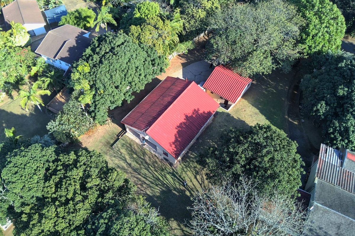 Aerial view of the property showing the main house with a red metal roof, surrounding garden, and mature trees on a spacious lot.