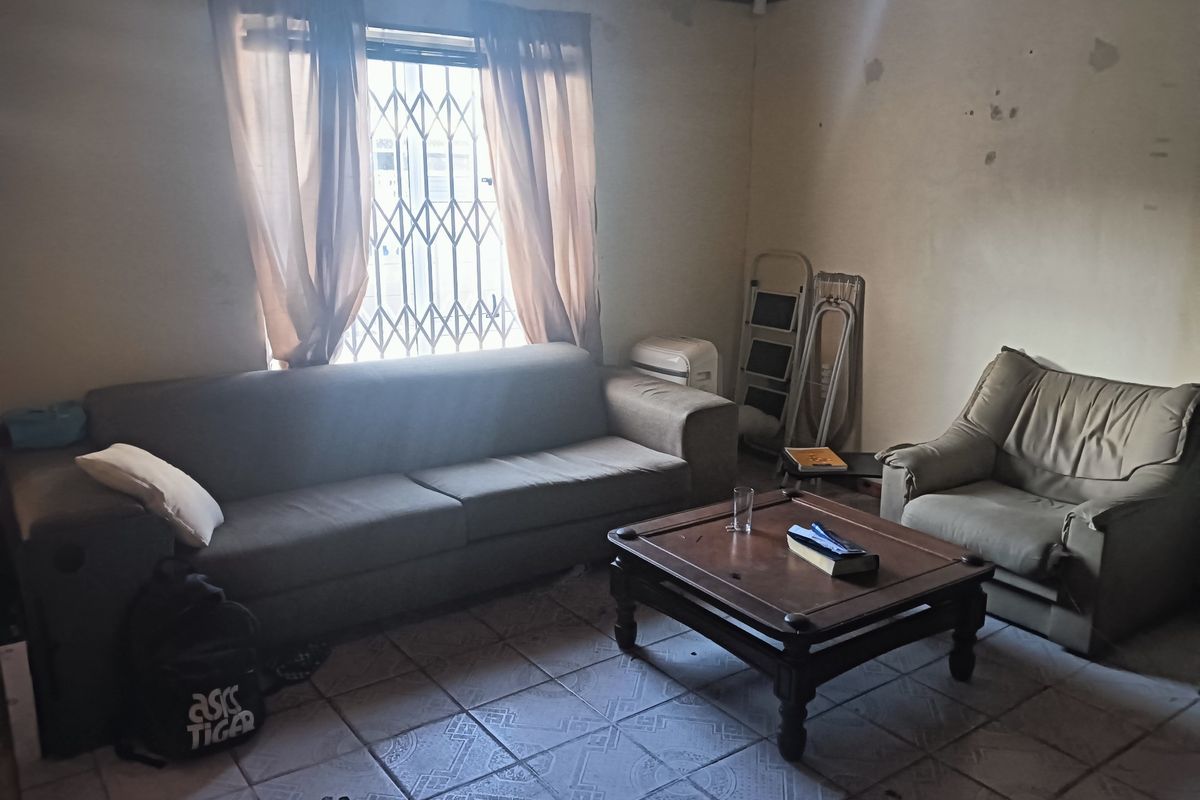 A living room featuring tiled flooring, a security gate on the window, and a timber ceiling, currently requiring cosmetic updates and wall repairs.