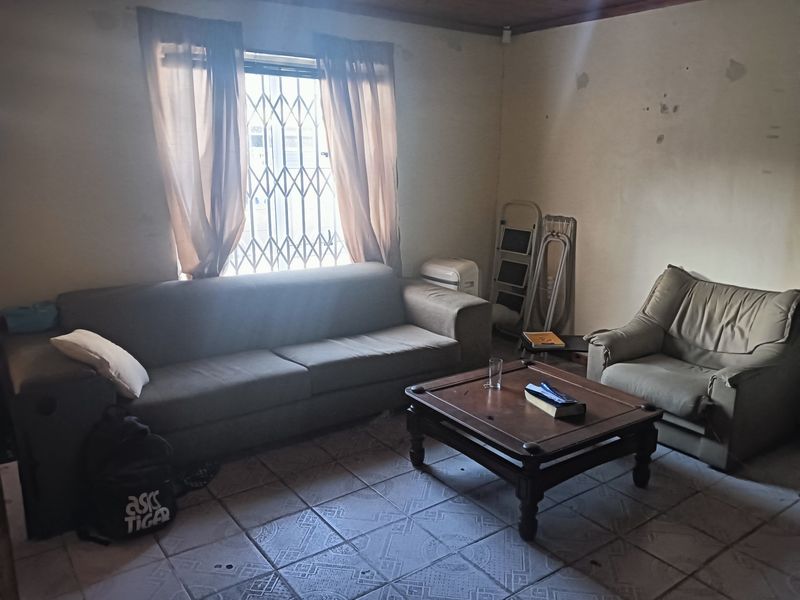 A living room featuring tiled flooring, a security gate on the window, and a timber ceiling, currently requiring cosmetic updates and wall repairs.