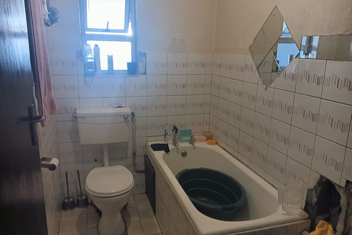 A dated bathroom featuring a standard bathtub and toilet with partially damaged wall tiling and exposed wall sections requiring repair and modernization.