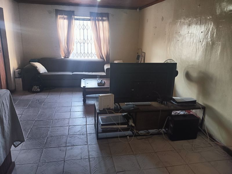 A living room featuring tiled flooring, a timber ceiling, and a window with a security gate, currently showing signs of wear on the walls.