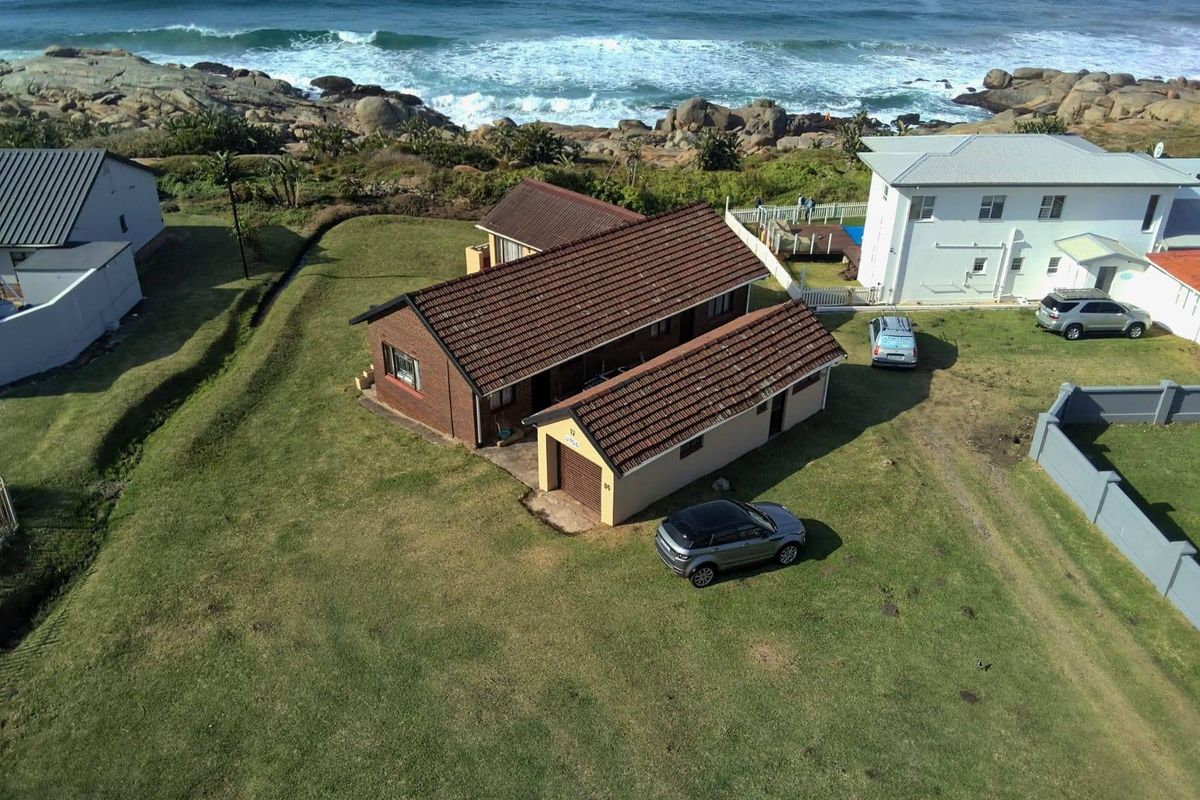 Aerial view of a coastal residential property featuring a brick house with a tiled roof, attached garage, and expansive lawn area overlooking the ocean.
