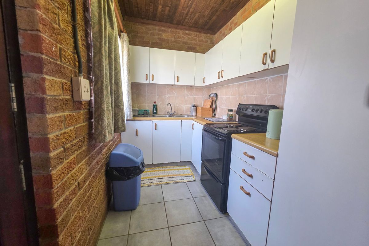 A compact kitchen featuring exposed brick walls, a timber ceiling, white cabinetry, and a freestanding stove with tiled splashback.