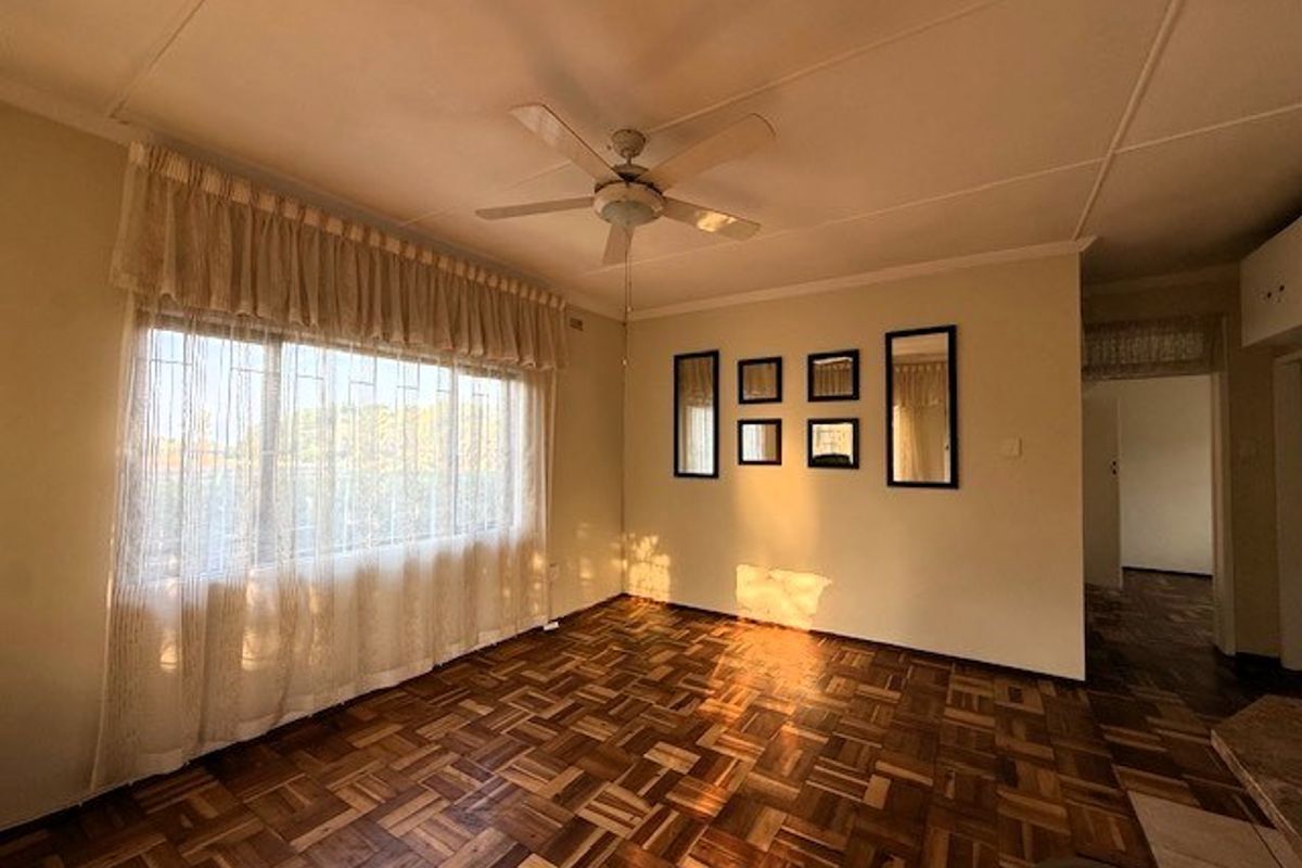 Spacious living room featuring traditional parquet flooring, a ceiling fan, and large windows with security bars, offering a bright and open living space.