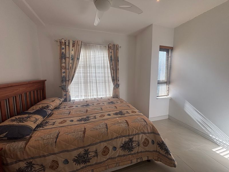 A well-lit bedroom featuring neutral tiled flooring, a ceiling fan, and dual window treatments including venetian blinds and curtain rods.