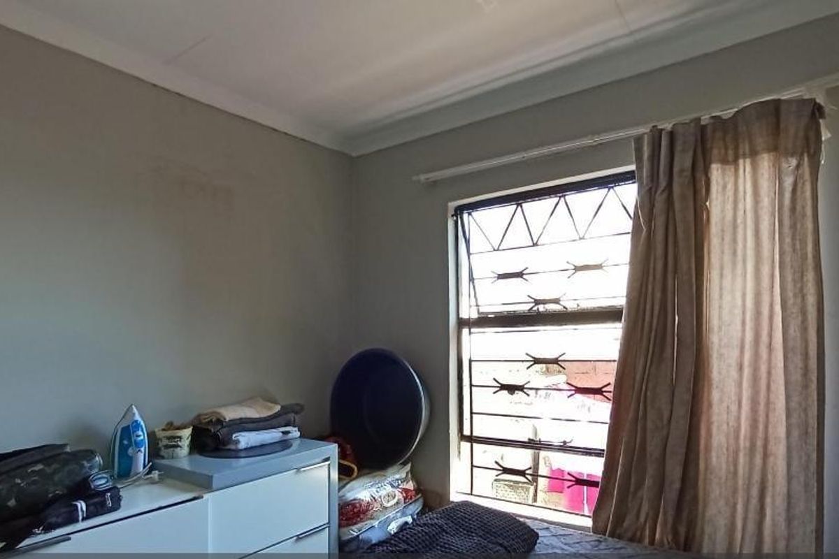 A bedroom featuring a window with security bars and a curtain rod, with visible wear on the wall surfaces.