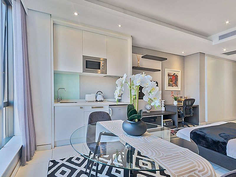 Modern open-plan apartment living space featuring a compact kitchenette with built-in cabinetry, integrated microwave, and sleek finishes, complemented by recessed lighting and ducted air conditioning.