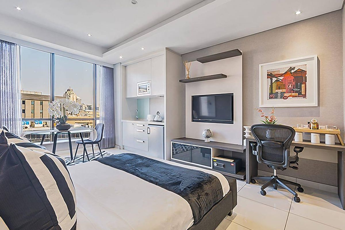 Modern bedroom featuring a compact built-in kitchenette, floor-to-ceiling windows with city views, recessed lighting, and sleek tiled flooring.